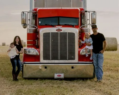 Mother, Father and daughters standing in front of their red Peterbilt 389 class 8 truck on a field with hay bales behind them