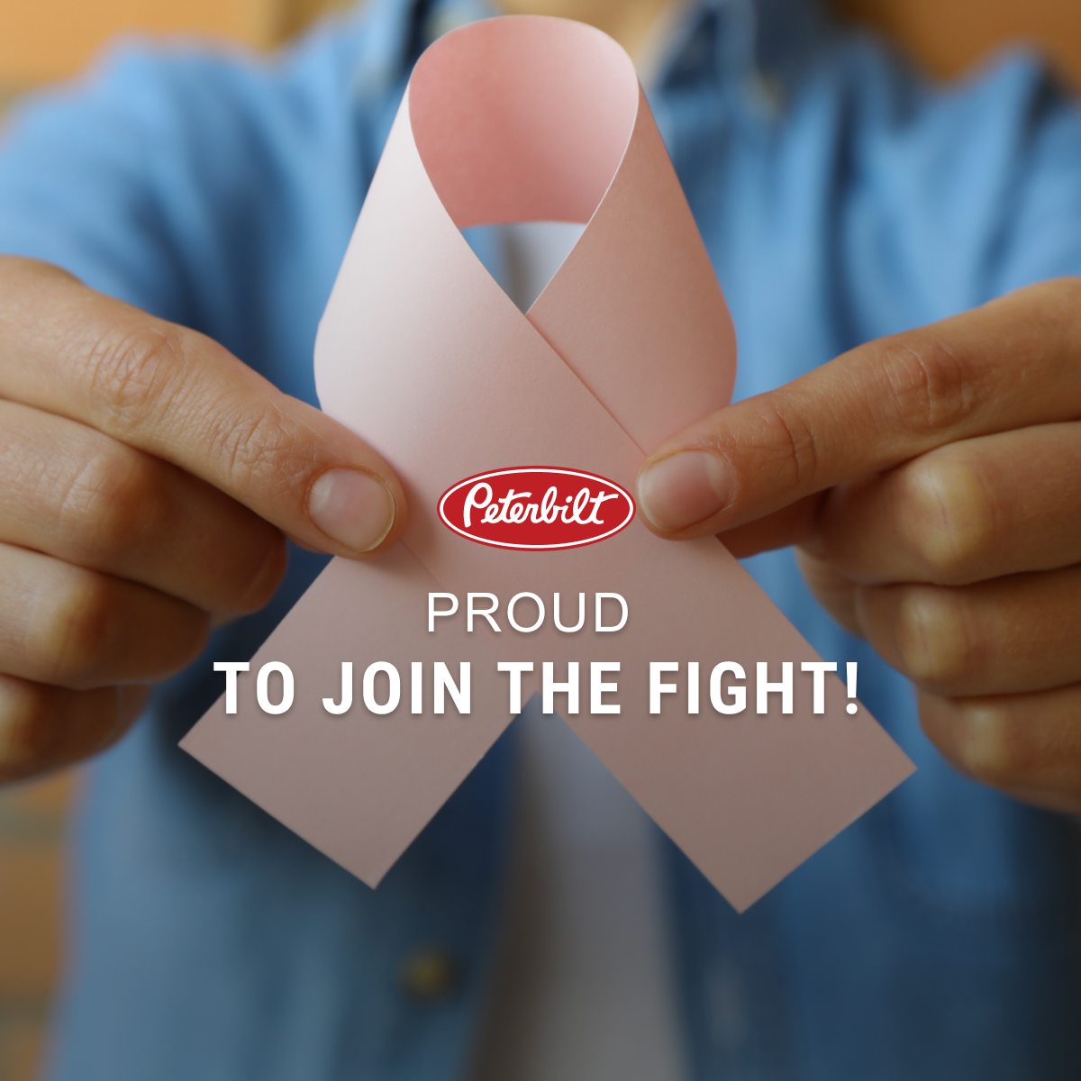 woman in blue shirt holding pink ribbon to the camera with Peterbilt logo and proud to join the fight type